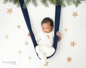 Baby in a white knit suit sits in a navy hanging swing with star decorations on a white backdrop.