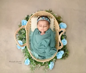 Baby sleeping peacefully in a woven basket, swaddled in teal fabric with a blue floral headband, surrounded by blue roses and greenery on a beige background.