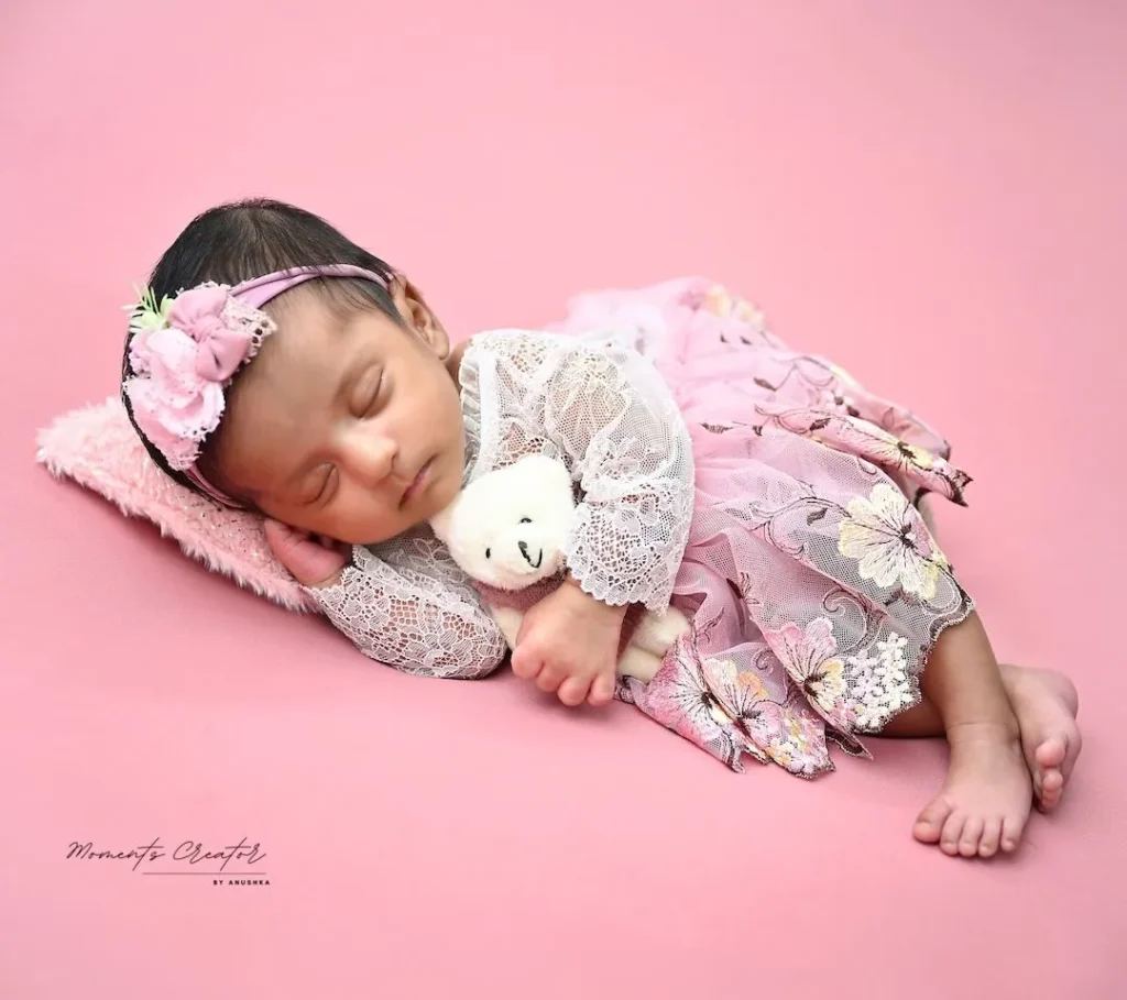 Newborn girl sleeping on a pink backdrop, wearing a lace dress and floral headband, cuddling a white teddy bear.