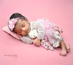 Newborn girl sleeping on a pink backdrop, wearing a lace dress and floral headband, cuddling a white teddy bear.